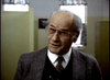 An older man with gray hair and glasses in a plaid blazer stands indoors before green partitions, resembling a character from classic psychological thrillers like "Murder By Reason of Insanity (1985)" on DVD.