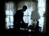 A man in a dark suit stands by a small table with a lamp and phone before sheer, sunlit curtains, capturing the suspenseful mood of "Murder By Reason of Insanity (1985) DVD.