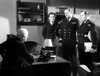 Four men in military uniforms stand in an office before a seated man, shelves and office supplies behind them—reminiscent of a scene from the classic WWII submarine drama, Morning Departure (1950) DVD.