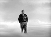 A man in a leather jacket and dress pants stands with hands in his pockets, gazing upward amid clouds, evoking the timeless style of Spencer Tracy as seen in the black-and-white classic "A Guy Named Joe (1943)" DVD.
