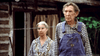 An elderly woman in a patterned blouse stands with an elderly man in plaid shirt and overalls outside a rustic cabin—a scene reminiscent of Empire of the Ants (1977) DVD, framed by curtains and lush greenery.