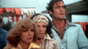 Three people stand close together outdoors, looking worried—like a scene from a cult sci-fi horror film. Colorful banners and an out-of-focus structure hint at a festive event. From the 1977 classic "Empire of the Ants," available now on DVD.