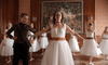 In an ornate room, a teacher guides young dancers in white dresses and colored sashes as they practice ballet positions with grace reminiscent of scenes from The Ugly Stepsister DVD, all before a large decorative tapestry.