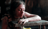 A young woman with blood on her face leans wearily on a rough wooden table, eyes closed, in a dim room—an evocative scene from The Ugly Stepsister DVD, ideal for collectors of rare films.