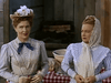 Two women in vintage attire stand outdoors by a table with a red-and-white checkered tablecloth, channeling the suspense of a Western thriller like the Silver Lode (1954) DVD, both wearing serious expressions before a wooden building.