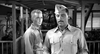 Two men in military uniforms stand in a covered outdoor walkway, evoking classic World War II drama as seen in Proud and Profane (1956) on DVD. The man in front has a mustache and cap, while the man behind him looks serious.