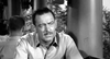 A man with short, dark hair and a mustache sits at a table in front of columns, wearing a light shirt. The black-and-white scene recalls classic films like Proud and Profane (1956), now available on DVD.