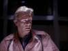A middle-aged person with short, gray-blonde hair and suspender straps stands in dim lighting, wearing a pink button-up over a blue shirt—ready to hunt for rare films like the Dead Space (1991) DVD, a classic sci-fi horror movie.