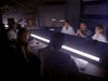 A group sits around a futuristic, U-shaped table in a dim room, discussing the rare Dead Space (1991) DVD. Papers and monitors are scattered on the table as a large screen glows in the background.