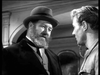Two men talk indoors: one bearded in a bowler hat and suit, the other clean-shaven in lighter clothes. The black-and-white scene recalls classic British POW film moments from the Seven Thunders (1957) DVD and other WWII movies.