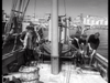 Fishermen sort their catch on a boat's deck, surrounded by baskets and gear. Across the harbor, buildings and boats under a clear sky evoke scenes from Seven Thunders (1957) DVD.