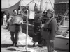 A stern soldier in uniform stands on a dock near fishing boats as two men walk by, capturing the tense mood of war in this old black-and-white scene from the Seven Thunders (1957) DVD.