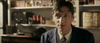 A man in a suit and tie with wavy hair sits indoors, smiling slightly. Shelves behind him display canned goods, bottles, and The Pardon (2013) DVD. A vintage fan rests on a table under warm, soft lighting.