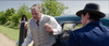 A man in a light vest and tie steps from a vintage car, looking worried, as seen in "The Pardon (2013) DVD." Nearby stands a man in a dark suit and hat, while a woman in a hat is visible among trees by the roadside.