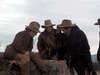 Four people in brown frontier clothing and wide-brimmed hats sit and kneel around a tree stump outdoors under a cloudy sky—reminiscent of scenes from the rare Winds of Autumn (1976) DVD.