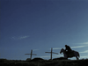 A person on a white horse sits by two wooden crosses on a grassy hill under a blue sky—evoking scenes from rare, classic films like "Winds of Autumn (1976)" now available on DVD.