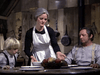 A woman with gray hair in a white apron serves food to a man and young blond boy at a rustic table, while the "Winds of Autumn (1976) DVD" plays softly in the cozy, old-fashioned kitchen.