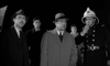 A man in a trench coat and fedora, a police officer, and others in suits gather under bright lights at night—a scene echoing classic British sci-fi horror as seen in The Quatermass Xperiment (1955) DVD.