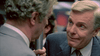 Two men in suits stand close together on a subway, radiating the suspense of a thriller. One faces the camera with a sly smirk reminiscent of Michael Caine in A Shock to the System (1990) DVD; the other is seen from behind.