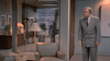 A man in a gray suit stands in an office near a green armchair and floor lamp, evoking the mood of the film "A Shock to the System (1990)" DVD. Through the glass wall, people are seen talking in the hallway outside.