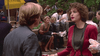 A woman in a red blazer holding a Diet Coke chats with a man in a suit outdoors, evoking Michael Caine’s sharp energy from A Shock to the System (1990) DVD, as others in business attire mingle near trees and a hot dog stand.
