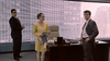 In a scene from "A Shock to the System (1990)" DVD, three people are in a modern office: a woman in yellow holding papers, and two men—one by the window and one at the desk, both set against large windows with blinds.
