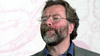 A middle-aged man with glasses and a graying beard looks slightly upward, dressed in a blue shirt and gray blazer against a pink and white backdrop—reminiscent of the Bloodlines (2010) DVD cover.