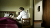 A man sits on a chair by a young girl in bed under a burgundy blanket in a softly lit room, gently watching over her—a quiet, caring scene reminiscent of moments from the "Bloodlines (2010) DVD.