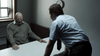 Two men are in a dim room. An older man sits tensely at a table as a younger man in office attire leans in to question him; a clock hangs on the wall—a scene echoing the intense moments found in the Bloodlines (2010) DVD.