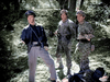 Three men in military uniforms stand in a wooded area—one wears a leather jacket and cap, the others are in camouflage. A fourth lies at their feet. Sunlight filters through the trees in this classic scene from "Fear and Desire" (1952) on DVD.