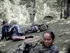 Three men in military-style clothing lie on the forest floor, one sitting up and facing the camera, while a rifleman stands in the background—a striking scene from "Fear and Desire" (1952), available as a rare DVD.