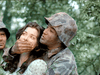 A tense scene reminiscent of a classic war movie unfolds in the woods: in Fear and Desire (1952), a soldier in camouflage restrains an anxious woman, covering her mouth, while another camouflaged figure is partially visible on the left.