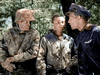 Three men in vintage military uniforms converse outdoors in dappled sunlight and trees—two wear camouflage, one sports a dark pilot jacket with aviator wings—evoking a scene from Fear and Desire (1952).