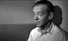 A middle-aged man with short hair sits against a wall in a light collared shirt, gazing downward thoughtfully. The black-and-white image evokes classic scenes like those in the "On The Beach (1959) DVD.