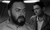 Two men feature in a black-and-white scene—one in uniform looks serious in the foreground, while another stands behind in a jacket and checked shirt. Experience this powerful moment on "On The Beach (1959)" DVD—a must-have for film enthusiasts.
