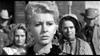 A close-up of a woman with curly blonde hair and large earrings, dressed in vintage style, evokes classic Westerns—reminiscent of "Terror in a Texas Town (1958) DVD" cover art—with other women blurred behind her in black and white.