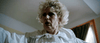 An older woman with curly blonde hair and a serious expression wears a white nightgown with a lace collar, standing indoors as light filters through the window—a scene of supernatural terror like that in Haunted Hotel (2021) DVD.
