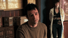 A man with short dark hair and a brown sweater looks concerned, while a woman in a light coat stands by cardboard boxes of "The Man from Earth (2007)" DVDs in a dimly lit room.