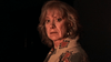 A woman with shoulder-length blond hair and a serious expression stands in dim lighting, her patterned red and gray jacket evoking the enigmatic style of "The Man from Earth (2007) DVD" as she glances over her shoulder at the camera.