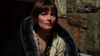 A woman with short brown hair and bangs sits indoors, clad in a blue fur-trimmed coat and layered necklaces, gazing off neutrally. The stone wall backdrop evokes a classic scene—like those in The Man from Earth (2007) DVD.