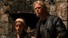 A man in a black leather jacket speaks animatedly while a woman with a pink headband sits beside him, both in front of a stone fireplace—echoing scenes from "The Man from Earth (2007)" DVD.