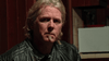 A middle-aged man with long blond hair and a goatee sits indoors in a black leather jacket, looking seriously at the camera as if discussing The Man from Earth (2007) DVD. Wooden panels and a white object appear in the background.