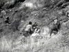 Two men in outdoor clothing crouch on a rocky hillside, partly hidden by dry grass, examining a person on the ground—a tense scene from the classic black-and-white Western, The Ride Back (1957) DVD starring Anthony Quinn.