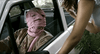 A person with their head wrapped in pink bandages sits in a car’s driver seat with the door open, looking up at someone outside—like a tense scene from the rare "Timecrimes (2007) DVD".