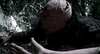 A figure in a rough brown sack crouches, reaching for motionless bare feet on the shadowy forest floor—evoking a scene from the Timecrimes (2007) DVD.