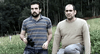 Two men are in a grassy field; one wears a striped polo holding binoculars, the other kneels beside him in a beige sweater. Tall trees line the background—an image reminiscent of the Timecrimes (2007) DVD.
