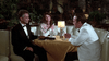 Three people in formal wear sit at a yellow table in a plush restaurant booth—a scene reminiscent of classic movies, as seen in the "Bogie (1980) DVD." A man in black tie faces a woman and another man in a white tuxedo, with drinks and a lamp on the table.
