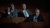 In an empty, dimly lit theater, three men in suits sit seriously focused—a scene reminiscent of a Humphrey Bogart biopic. The man on the left holds a cigarette. Product: Bogie (1980) DVD.