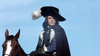 A person dressed like Richard Chamberlain in "The Man in the Iron Mask (1977) DVD," with a large black hat, white plume, cloak, and gloves, sits on a brown horse under a clear blue sky.