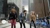 Three women on a city street near Citi Bike bicycles look surprised—as if spotting a Sharknado 2: The Second One (2014) DVD in a shop window. Tall buildings and cloudy skies complete the urban scene.